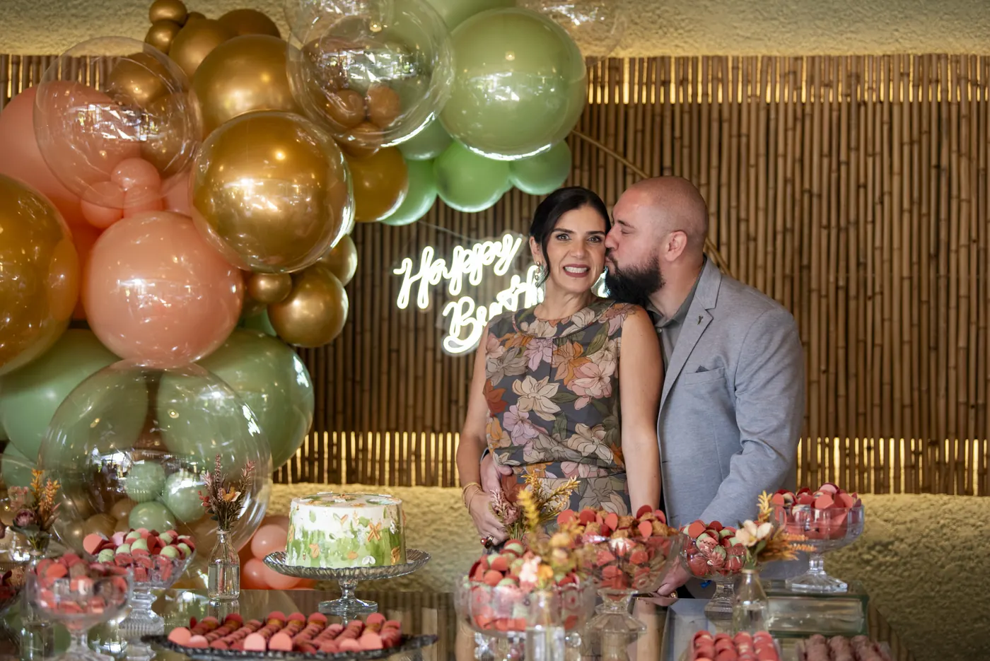 Casal na mesa de doces com arco de balões dourados e verdes diante da parede de bambu