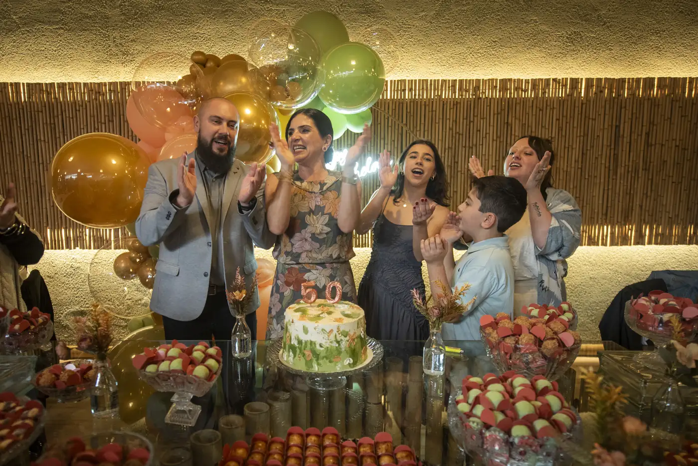 Mesa do bolo com doces e balões e convidados aplaudindo a celebração