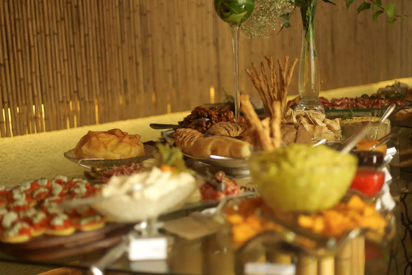 Buffet de aperitivos com arranjo floral alto diante da parede de bambu dourada