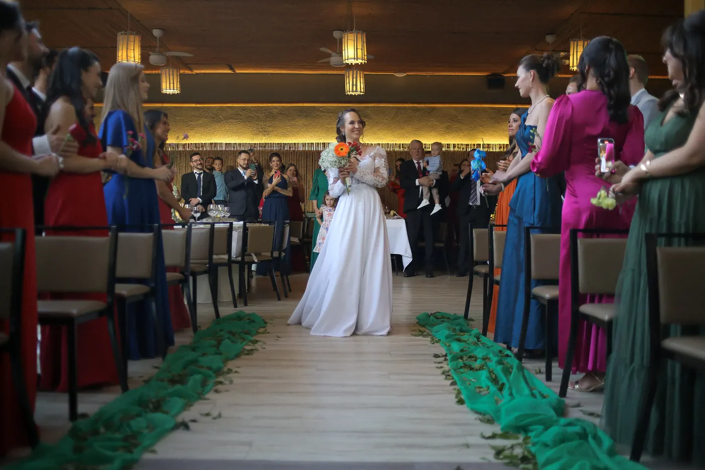 Noiva no corredor com madrinhas de vestidos coloridos e pétalas verdes no chão