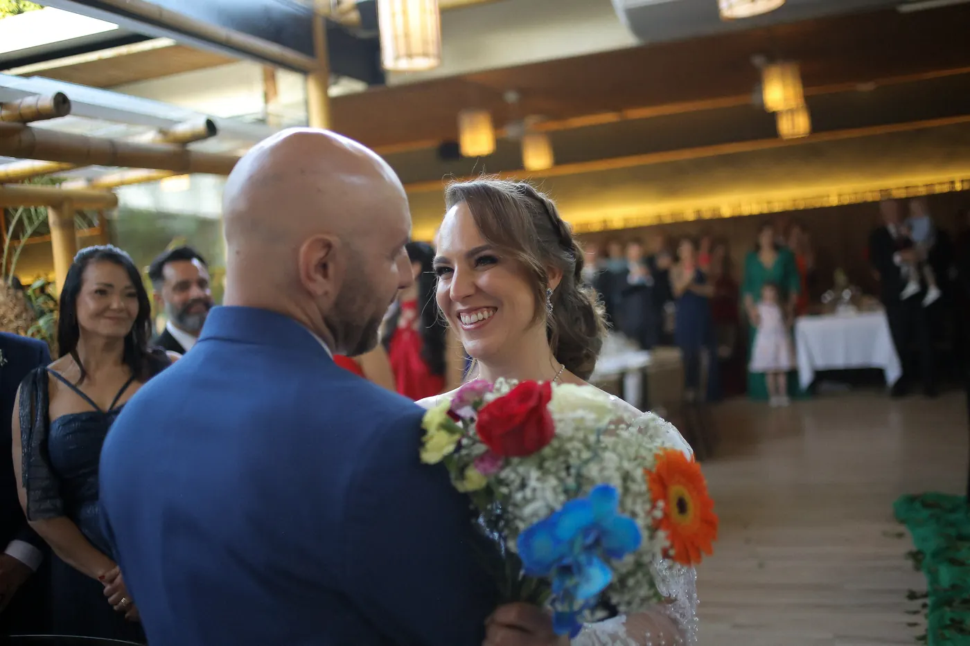 Noivos sorrindo durante a cerimônia com buquê colorido e convidados ao fundo